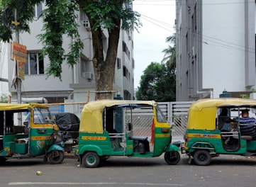 Three colorful auto rickshaws are parked in a row on the side of a street. Each has a green and yellow color scheme with decorative elements. The surroundings include tall residential buildings and lush green trees. A sign indicating a hostel is visible on the left side.