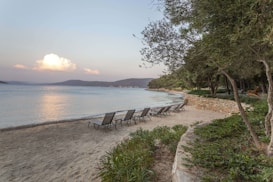 Several lounge chairs are arranged on a sandy beach next to a calm body of water. The beach is lined with lush trees and greenery. In the background, there are hills and mountains, under a sky with a prominent cloud formation. A stone wall runs parallel to the water, bordering the greenery.