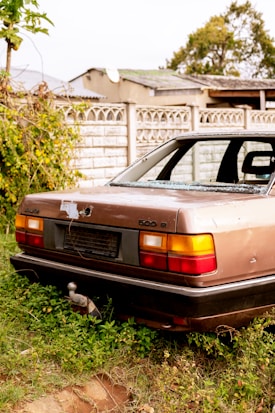 A worn-out and abandoned car with broken windows is parked on overgrown grass. The background features a white brick wall and a few trees. The car has a light brown color with a faded and damaged exterior, including a missing license plate and visible rust.