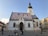 A historic church with a distinct colorful tiled roof featuring two coats of arms. The building showcases Gothic architectural elements and has a tall bell tower. In front of the church, two people in traditional attire stand beside a cart in a cobblestone plaza.