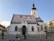 A historic church with a distinct colorful tiled roof featuring two coats of arms. The building showcases Gothic architectural elements and has a tall bell tower. In front of the church, two people in traditional attire stand beside a cart in a cobblestone plaza.