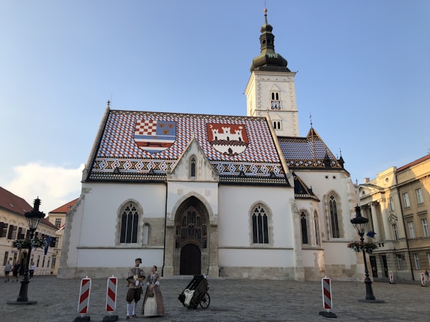 A historic church with a distinct colorful tiled roof featuring two coats of arms. The building showcases Gothic architectural elements and has a tall bell tower. In front of the church, two people in traditional attire stand beside a cart in a cobblestone plaza.