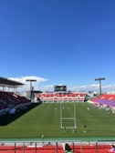 A large sports stadium with red and blue seating overlooks a green field with a goalpost at the center. A few people are scattered across the field, with some tents and a gathering of individuals on the right side. The sky is clear and blue with some clouds visible in the distance. A large screen or scoreboard is situated at the far end of the field.