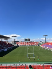 A large sports stadium with red and blue seating overlooks a green field with a goalpost at the center. A few people are scattered across the field, with some tents and a gathering of individuals on the right side. The sky is clear and blue with some clouds visible in the distance. A large screen or scoreboard is situated at the far end of the field.