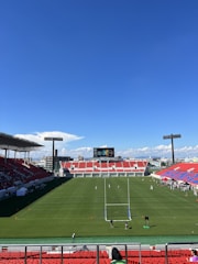 A large sports stadium with red and blue seating overlooks a green field with a goalpost at the center. A few people are scattered across the field, with some tents and a gathering of individuals on the right side. The sky is clear and blue with some clouds visible in the distance. A large screen or scoreboard is situated at the far end of the field.