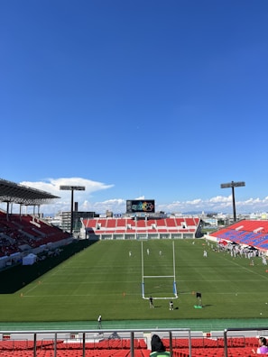 A large sports stadium with red and blue seating overlooks a green field with a goalpost at the center. A few people are scattered across the field, with some tents and a gathering of individuals on the right side. The sky is clear and blue with some clouds visible in the distance. A large screen or scoreboard is situated at the far end of the field.