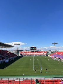 A large sports stadium with red and blue seating overlooks a green field with a goalpost at the center. A few people are scattered across the field, with some tents and a gathering of individuals on the right side. The sky is clear and blue with some clouds visible in the distance. A large screen or scoreboard is situated at the far end of the field.