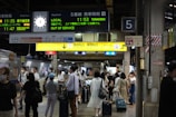 A busy train station platform with a diverse group of people, some wearing masks, and various signs in English and Japanese. Passengers are standing with luggage, waiting or moving towards an exit. Electronic displays show train schedules and destinations. The atmosphere is active and bustling.
