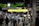 A busy train station platform with a diverse group of people, some wearing masks, and various signs in English and Japanese. Passengers are standing with luggage, waiting or moving towards an exit. Electronic displays show train schedules and destinations. The atmosphere is active and bustling.