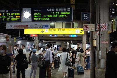 A busy train station platform with a diverse group of people, some wearing masks, and various signs in English and Japanese. Passengers are standing with luggage, waiting or moving towards an exit. Electronic displays show train schedules and destinations. The atmosphere is active and bustling.