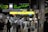 A busy train station platform with a diverse group of people, some wearing masks, and various signs in English and Japanese. Passengers are standing with luggage, waiting or moving towards an exit. Electronic displays show train schedules and destinations. The atmosphere is active and bustling.