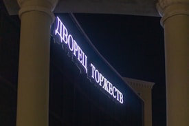 Illuminated sign with Cyrillic text on a building fa&ccedil;ade, framed by two large columns and set against a dark night sky.