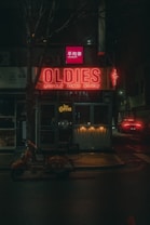 A dimly lit urban street scene featuring a neon sign displaying the word 'OLDIES' in red light. Below the sign, there's an entrance to a venue that seems to offer various tacos and drinks. A vintage orange scooter is parked on the sidewalk in front of the establishment. A red car with its taillights on is visible driving down the street in the distance.