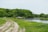 A serene natural landscape with a dirt path winding through lush green grass alongside a calm river. Dense trees, rich in green foliage, create a wooded backdrop. There are a few cars parked near the trees, suggesting a rural or recreational setting.