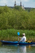 A person in a white long-sleeve shirt and a hat is kayaking on a river. The kayak is blue with some equipment, including a yellow bag, on board. There is dense greenery along the riverbank, and in the background, there are tall trees and what appears to be a church with golden domes.