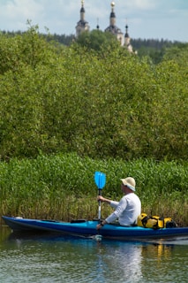A person in a white long-sleeve shirt and a hat is kayaking on a river. The kayak is blue with some equipment, including a yellow bag, on board. There is dense greenery along the riverbank, and in the background, there are tall trees and what appears to be a church with golden domes.