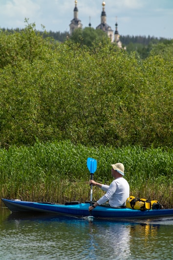 A person in a white long-sleeve shirt and a hat is kayaking on a river. The kayak is blue with some equipment, including a yellow bag, on board. There is dense greenery along the riverbank, and in the background, there are tall trees and what appears to be a church with golden domes.