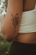 A close-up of a person's arm with a delicate floral tattoo in black ink. The person is wearing a white sleeveless top, and the background is softly blurred with hints of greenery.