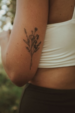 A close-up of a person's arm with a delicate floral tattoo in black ink. The person is wearing a white sleeveless top, and the background is softly blurred with hints of greenery.