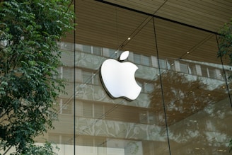 A large apple logo is displayed prominently on the glass facade of a modern building, surrounded by reflections of trees and the urban environment. The facade features a wooden ceiling, and the scene conveys a blend of nature and modern architecture.