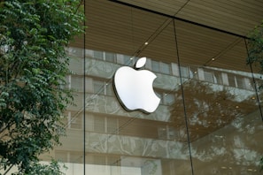A large apple logo is displayed prominently on the glass facade of a modern building, surrounded by reflections of trees and the urban environment. The facade features a wooden ceiling, and the scene conveys a blend of nature and modern architecture.