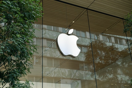 A large apple logo is displayed prominently on the glass facade of a modern building, surrounded by reflections of trees and the urban environment. The facade features a wooden ceiling, and the scene conveys a blend of nature and modern architecture.