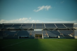 Empty stadium with blue bleachers, green grass field, and a clear blue sky. The stadium is large and expansive, featuring a scoreboard and a goalpost in the center. The seating is symmetric, and the sign 'Camping World' is visible.