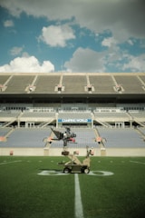 A large football stadium with empty tiered seating under a blue sky with scattered clouds. In the foreground, a camera rig on wheels is positioned on the grass field near the 50-yard line. The stadium has a 'Camping World' sign above the seating.