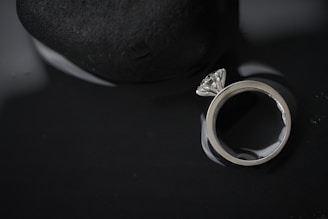 A diamond ring with a large clear stone is placed next to a smooth, dark stone. The background is dark, and the ring is highly reflective, creating a dramatic contrast between the bright metal and the dark surroundings.