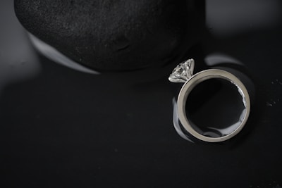 A diamond ring with a large clear stone is placed next to a smooth, dark stone. The background is dark, and the ring is highly reflective, creating a dramatic contrast between the bright metal and the dark surroundings.