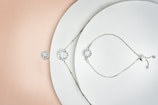 A delicately arranged collection of jewelry pieces, including a bracelet and a necklace, both featuring circular, diamond-like stones. The items are set against a minimalistic white background with a subtle gradient transitioning to a soft peach color. The jewelry pieces are metallic with small intricate details and clasps.