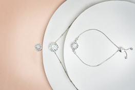A delicately arranged collection of jewelry pieces, including a bracelet and a necklace, both featuring circular, diamond-like stones. The items are set against a minimalistic white background with a subtle gradient transitioning to a soft peach color. The jewelry pieces are metallic with small intricate details and clasps.