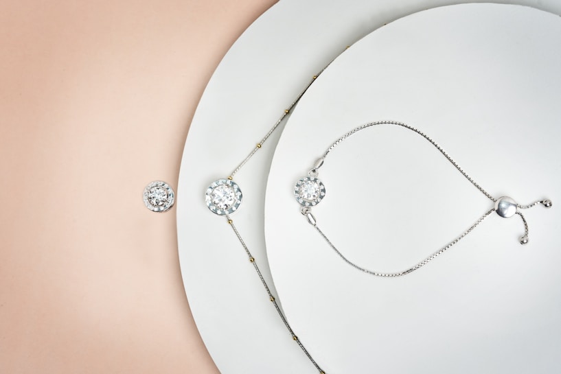 A delicately arranged collection of jewelry pieces, including a bracelet and a necklace, both featuring circular, diamond-like stones. The items are set against a minimalistic white background with a subtle gradient transitioning to a soft peach color. The jewelry pieces are metallic with small intricate details and clasps.