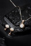 Elegant jewelry pieces, including a gold pendant with a large diamond and matching earrings, displayed on a wet, glossy black stone surface with water droplets.