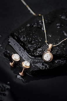 Elegant jewelry pieces, including a gold pendant with a large diamond and matching earrings, displayed on a wet, glossy black stone surface with water droplets.