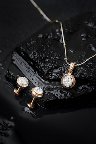 Elegant jewelry pieces, including a gold pendant with a large diamond and matching earrings, displayed on a wet, glossy black stone surface with water droplets.