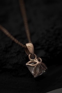 A close-up view of a gold pendant with an intricate floral design hanging from a delicate chain. The pendant rests on a dark textured surface, creating a contrast that highlights the shine and details of the jewelry.