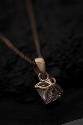 A close-up view of a gold pendant with an intricate floral design hanging from a delicate chain. The pendant rests on a dark textured surface, creating a contrast that highlights the shine and details of the jewelry.