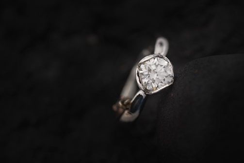 A close-up image of a diamond ring resting on a dark, textured surface. The ring features a prominent, round-cut diamond set in a simple, elegant band, with the dark background providing contrast to the brilliance of the gemstone.