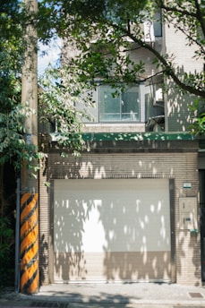A closed garage door is surrounded by a brick wall with a window section above it. There is an abundance of green leaves from nearby trees casting shadows on the structures. A utility pole with yellow and black diagonal stripes stands to the left.