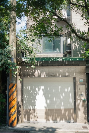 A closed garage door is surrounded by a brick wall with a window section above it. There is an abundance of green leaves from nearby trees casting shadows on the structures. A utility pole with yellow and black diagonal stripes stands to the left.