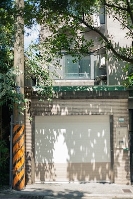 A closed garage door is surrounded by a brick wall with a window section above it. There is an abundance of green leaves from nearby trees casting shadows on the structures. A utility pole with yellow and black diagonal stripes stands to the left.