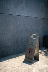 A worn, A-frame sign with the text 'NO PARKING' and a circular red prohibition symbol stands on a sunlit concrete surface against a plain, dark gray wall.