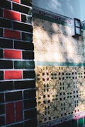 A wall with intricately designed ceramic tiles, featuring a variety of patterns and colors. On the left, darker brick tiles with green and red colors, while the right shows lighter tiles with floral motifs and geometric shapes in shades of beige, green, and red. Light and shadow create a dynamic play across the surface, adding depth to the textures.