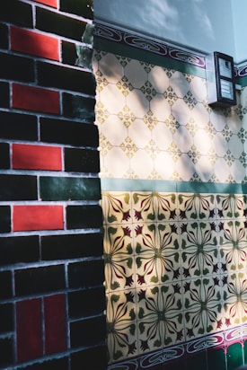 A wall with intricately designed ceramic tiles, featuring a variety of patterns and colors. On the left, darker brick tiles with green and red colors, while the right shows lighter tiles with floral motifs and geometric shapes in shades of beige, green, and red. Light and shadow create a dynamic play across the surface, adding depth to the textures.