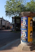 A street scene featuring a cylindrical poster column covered with various posters. The posters advertise an event or performance, with prominent bold text and graphics. The surrounding area includes charming traditional European-style buildings, a tree with green leaves, and a paved road. The scene is well-lit, indicating daytime and clear weather.