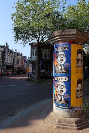 A street scene featuring a cylindrical poster column covered with various posters. The posters advertise an event or performance, with prominent bold text and graphics. The surrounding area includes charming traditional European-style buildings, a tree with green leaves, and a paved road. The scene is well-lit, indicating daytime and clear weather.