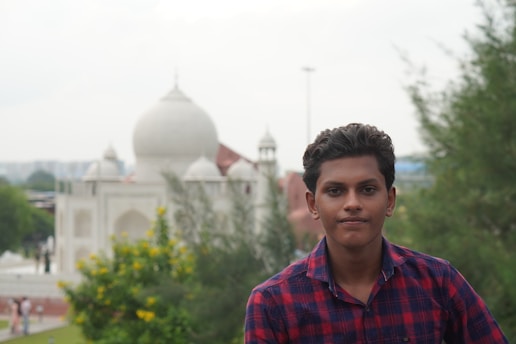 A young person wearing a red and blue checkered shirt is in the foreground. In the background, there is a large white building with domes reminiscent of Mughal architecture, surrounded by greenery.