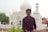 A young man in a checkered shirt stands in front of a large white building with ornate detailing and a dome structure, resembling traditional Mughal architecture. The scene includes other people in the background walking and taking photos, with greenery in the foreground.