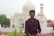 A young man in a checkered shirt stands in front of a large white building with ornate detailing and a dome structure, resembling traditional Mughal architecture. The scene includes other people in the background walking and taking photos, with greenery in the foreground.